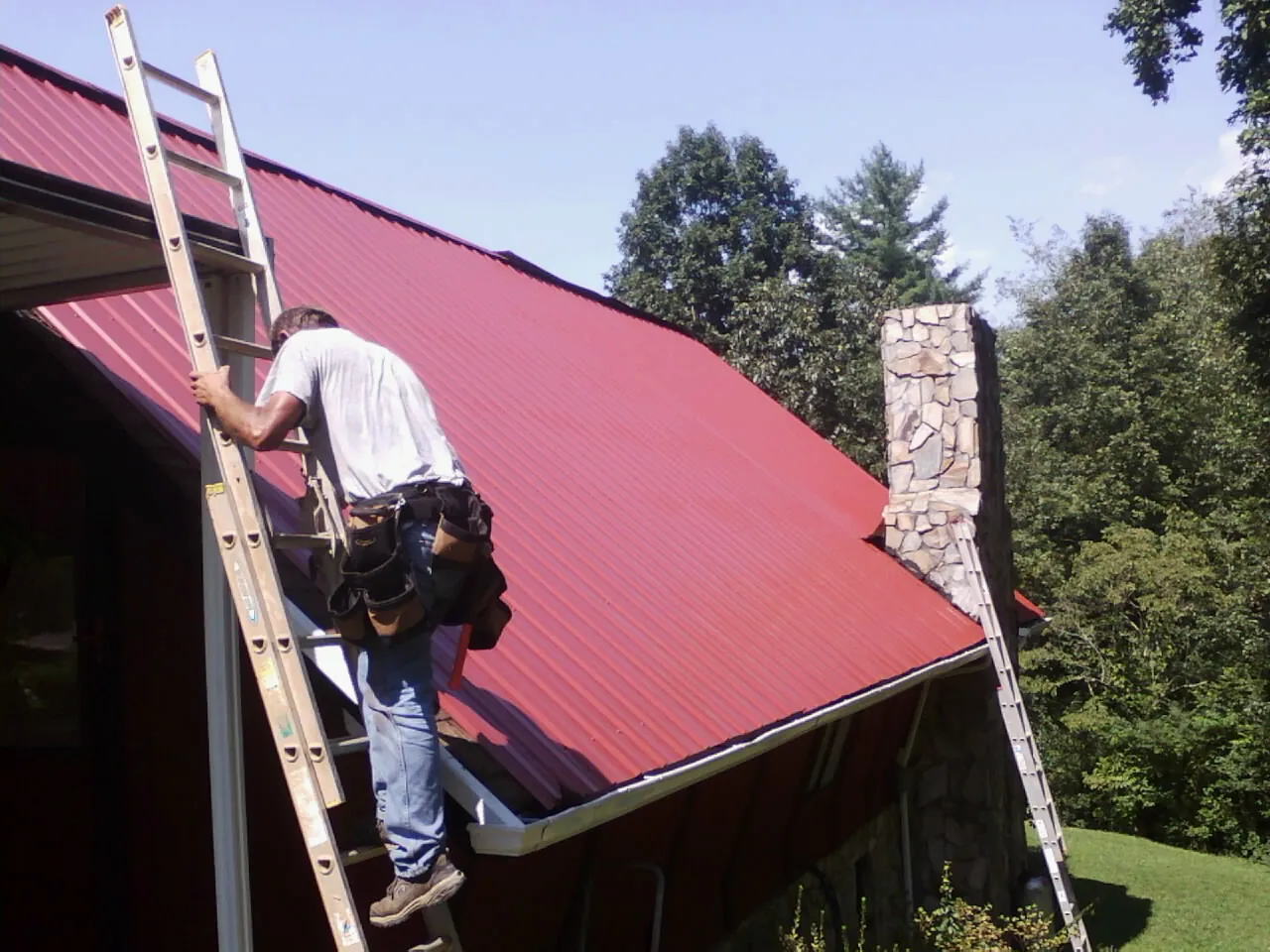 Roofer on a ladder beside a red metal roof and stone chimney — Soffit & Fascia Repair by Roofing Brothers in Derwood
